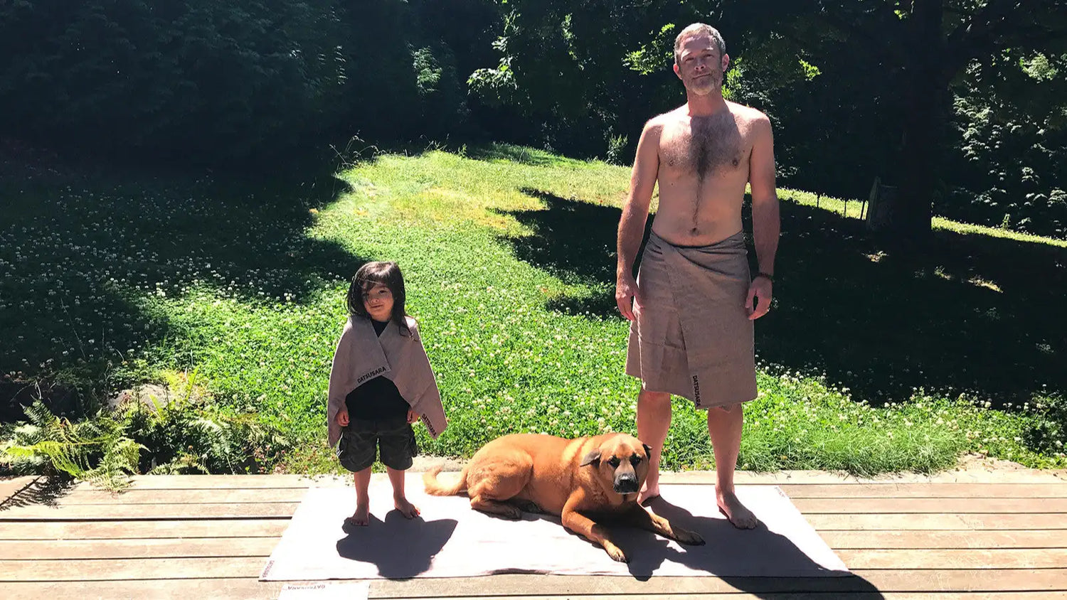 Man, child, and dog standing on a wooden deck with grass and trees in the background, wearing towels by Datsusara