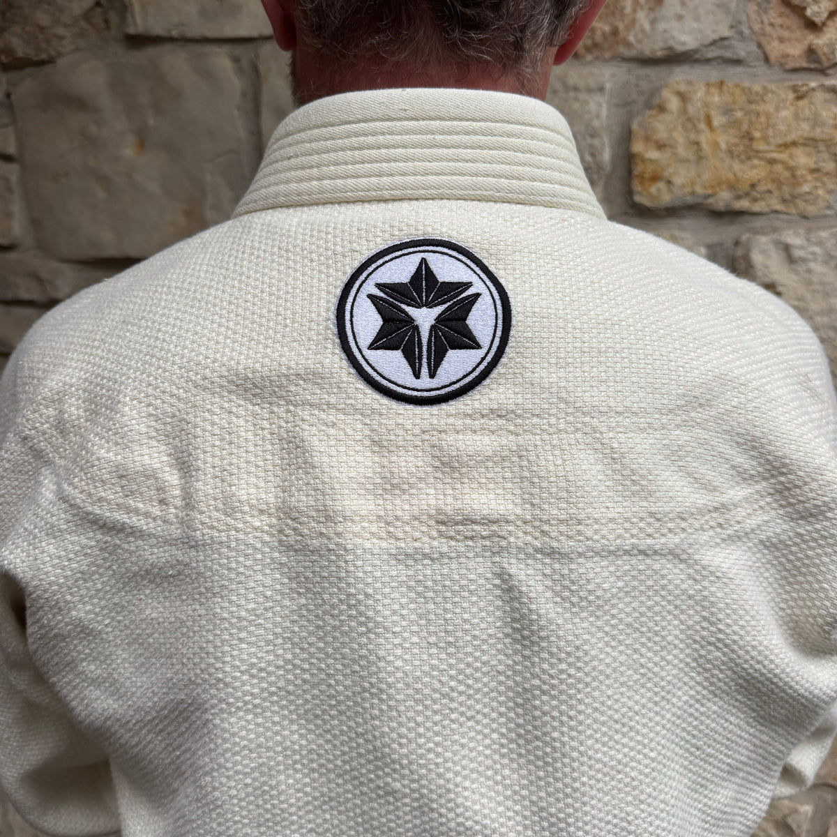 Person wearing a beige martial arts uniform with a black and white emblem on the back against a stone wall.