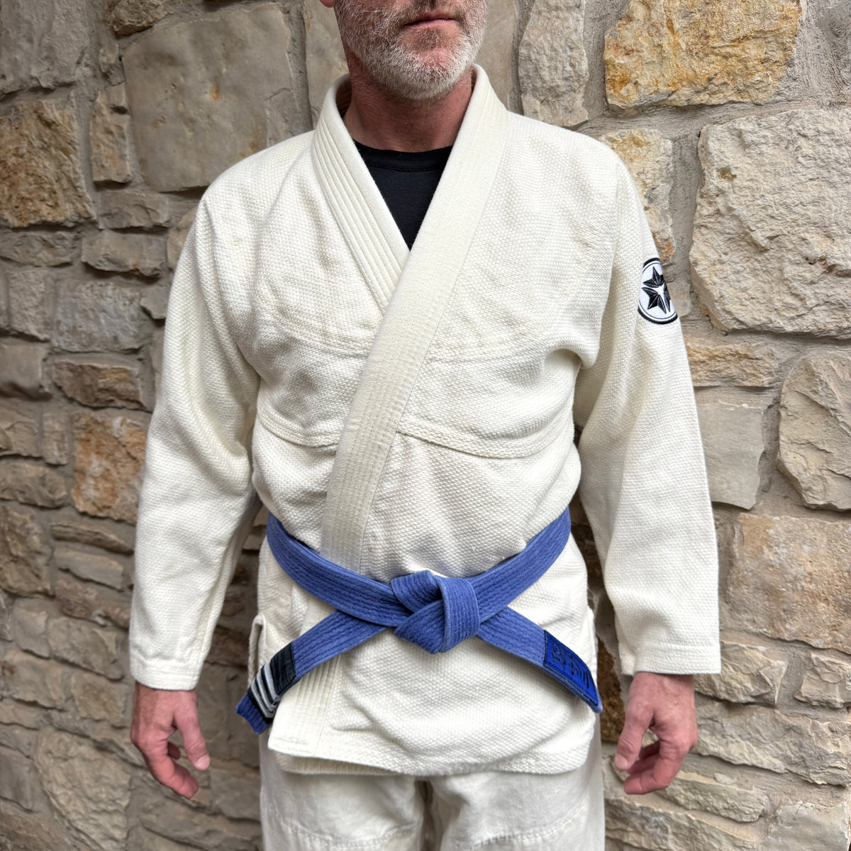 Man wearing a white martial arts uniform with a blue belt against a stone wall.