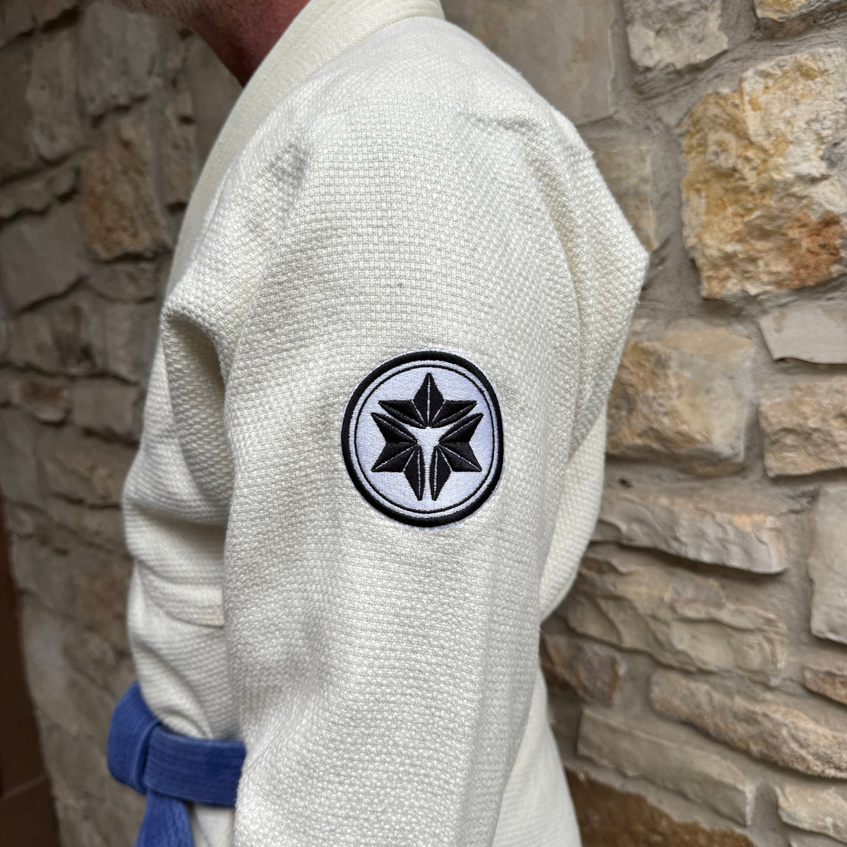 Person wearing a white martial arts gi with a black and white emblem on the sleeve, standing against a stone wall.