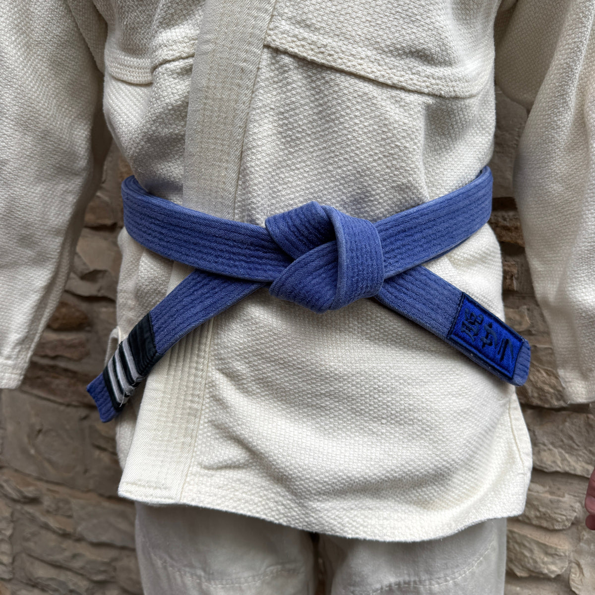Person wearing a white martial arts uniform with a blue belt against a stone wall background