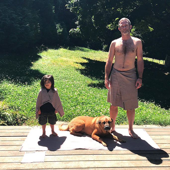 An image showing a hemp towel being used for yoga on a grassy outdoor area with a person standing next to it and a dog lying down on the towel.